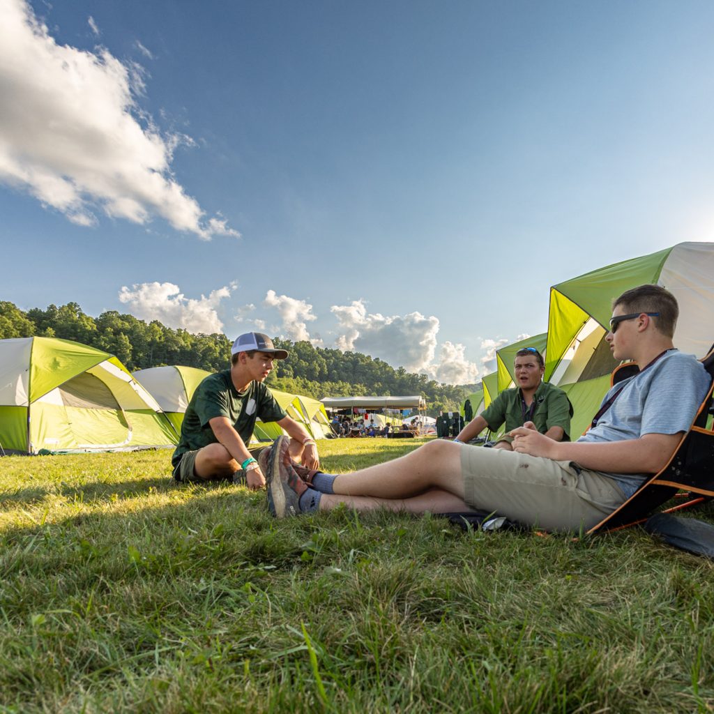 Jack Harkins, Riley Dee, and Wiley Walrod of the Mount Baker Council chat as the sun sets during the 2023 National Scout Jamboree at The Summit Bechtel Reserve in Mount Hope, West Virginia. (BSA Photo by Dan Glass) rows of yellow green tents..**********Beginning of Shooting Data Section**********.20230722-18-48-40-85-DG     date - 7/22/23     time - 6:48:40 PM